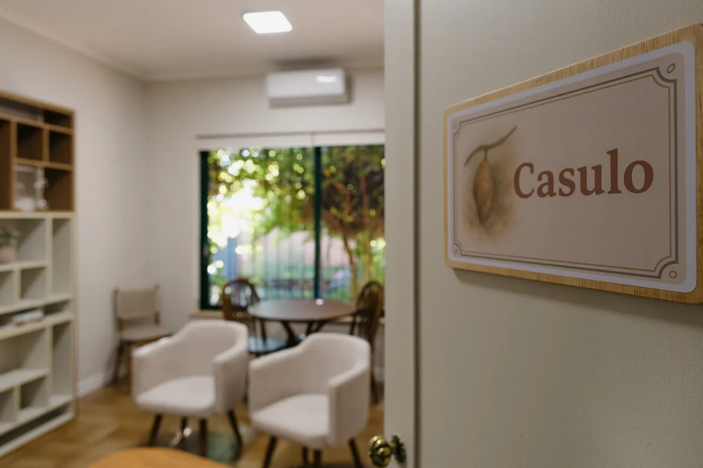 Porta com a placa Casulo aberta para uma sala com poltronas e janela para o jardim