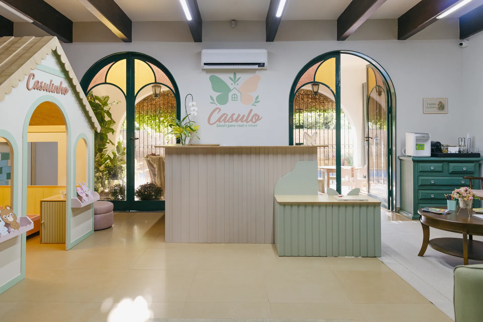 Recepção do Espaço Casulo, com portas em arco, balcão verde sálvia e a área infantil ao fundo